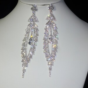 May include: A pair of dazzling, silver-toned chandelier earrings. Each earring features a geometric top with a cascade of sparkling, clear crystals. The crystals catch the light, creating a brilliant display of sparkle and shine. These earrings are a statement piece.