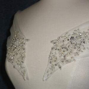 May include: Close-up of a white wedding dress with a beaded embellishment on the shoulder straps. The embellishment is made of small, clear crystals and is arranged in a floral pattern.