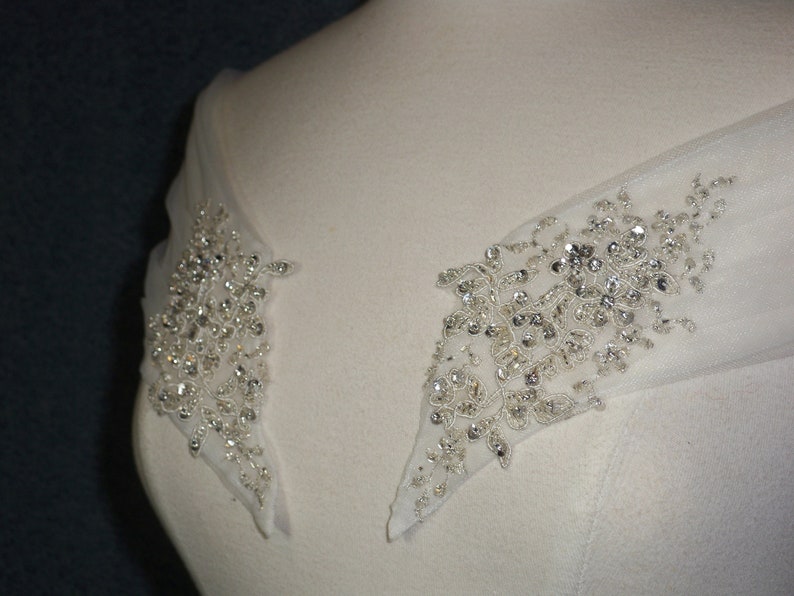 May include: Close-up of a white wedding dress with a beaded embellishment on the shoulder straps. The embellishment is made of small, clear crystals and is arranged in a floral pattern.