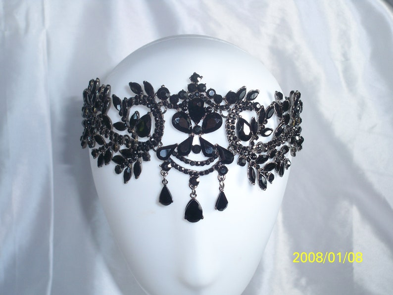 May include: A black, ornate headpiece with a symmetrical design. The headpiece features various shapes of black gemstones and dangling teardrop-shaped pendants. It is displayed on a white mannequin head against a white background.