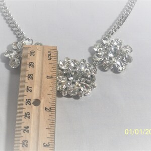 May include: A silver chain necklace with three large, clear rhinestone clusters. The center cluster is rectangular and the two side clusters are flower-shaped. The necklace is measured with a ruler, showing it is about 1 inch wide.