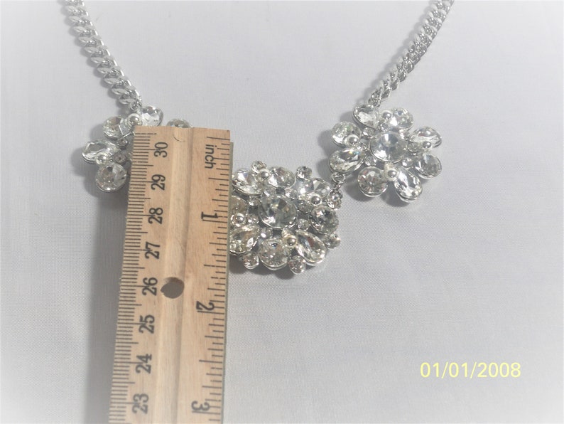 May include: A silver chain necklace with three large, clear rhinestone clusters. The center cluster is rectangular and the two side clusters are flower-shaped. The necklace is measured with a ruler, showing it is about 1 inch wide.