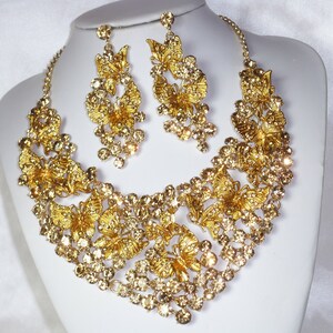 Puede incluir: Un conjunto de collar y pendientes dorados. El collar y los pendientes presentan un dise&ntilde;o de mariposa con cristales claros y brillantes. Las joyas se exhiben sobre un busto de maniqu&iacute; blanco sobre un fondo blanco.