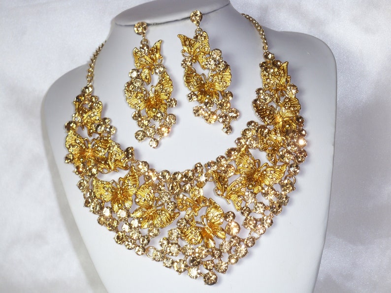 Puede incluir: Un conjunto de collar y pendientes dorados. El collar y los pendientes presentan un dise&ntilde;o de mariposa con cristales claros y brillantes. Las joyas se exhiben sobre un busto de maniqu&iacute; blanco sobre un fondo blanco.