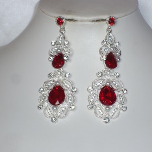 May include: A pair of silver-toned chandelier earrings with large, teardrop-shaped, red gemstones. The earrings feature intricate filigree detailing and smaller clear crystals. The earrings are displayed on a white mannequin head.