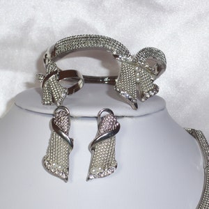 May include: A silver-toned jewelry set featuring a bracelet, ring, earrings, and necklace. The pieces are adorned with small, sparkling crystals and faux pearls. The bracelet and ring have a bow-like design, while the earrings and necklace have a cascading shape.