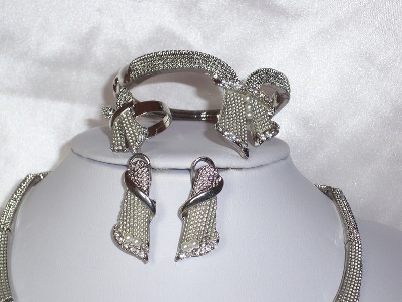 May include: A silver-toned jewelry set featuring a bracelet, ring, earrings, and necklace. The pieces are adorned with small, sparkling crystals and faux pearls. The bracelet and ring have a bow-like design, while the earrings and necklace have a cascading shape.
