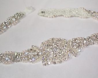 May include: A white bridal sash with a beaded design featuring clear rhinestones and pearls.