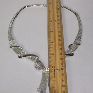 May include: Silver-toned necklace with a textured mesh design and a decorative pendant. The necklace features a rhinestone-studded accent and measures approximately 10 inches in length.