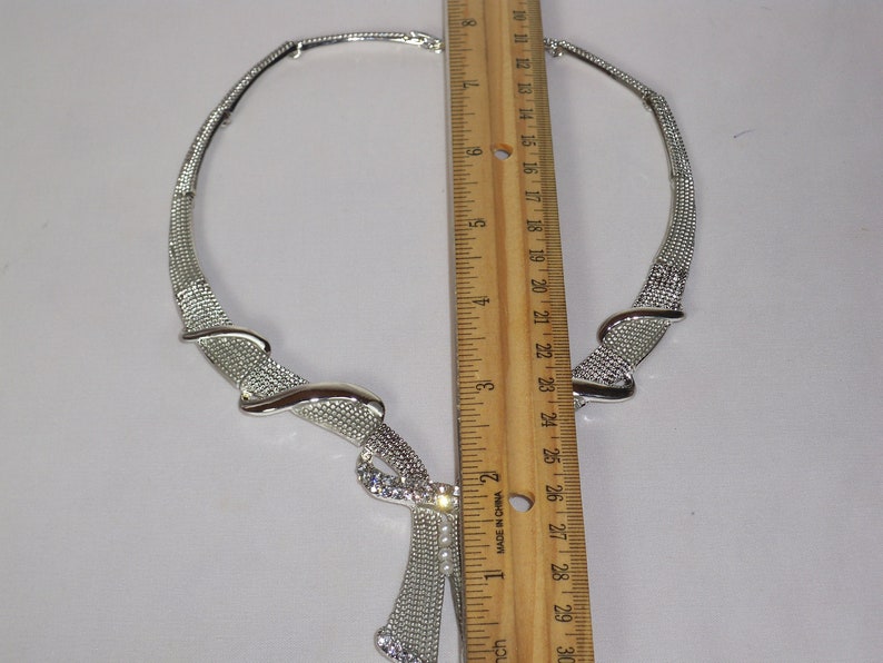 May include: Silver-toned necklace with a textured mesh design and a decorative pendant. The necklace features a rhinestone-studded accent and measures approximately 10 inches in length.