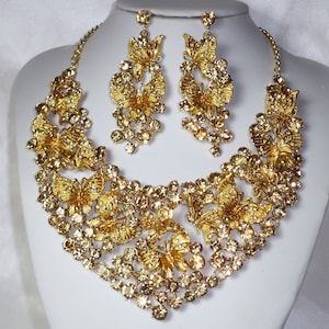 Puede incluir: Un conjunto de collar y pendientes dorados. El collar presenta intrincados dise&ntilde;os de mariposas y est&aacute; adornado con cristales brillantes. Los pendientes a juego tambi&eacute;n muestran motivos de mariposas y cristales colgantes.