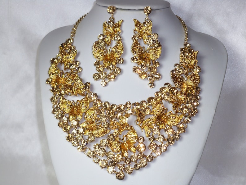 Puede incluir: Un conjunto de collar y pendientes dorados. El collar presenta intrincados dise&ntilde;os de mariposas y est&aacute; adornado con cristales brillantes. Los pendientes a juego tambi&eacute;n muestran motivos de mariposas y cristales colgantes.