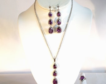 May include: A silver necklace and earrings set with three teardrop-shaped purple stones. The necklace has a pendant with three stones and the earrings have three stones each.