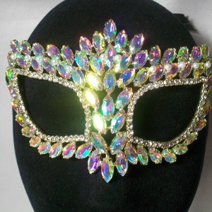 May include: A masquerade mask with iridescent rhinestones and faux pearls. The mask is decorated with a gold-tone metal frame and has a black velvet backing.