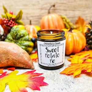 May include: A brown glass candle jar with a white label that reads "Chinesha's Closet Sweet Potato Pie". The candle is surrounded by fall-themed decor, including pumpkins, gourds, and leaves.