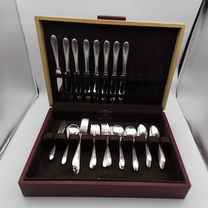 May include: A silver flatware set in a wooden case. The set includes knives, forks, and spoons, neatly arranged in a dark brown velvet-lined interior. The case is open, revealing the silverware's polished finish.