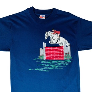May include: A navy blue t-shirt with a graphic of a white horse jumping over a brick wall. The horse is being ridden by a person wearing a red jacket and white pants. The graphic is surrounded by green grass and red flowers.