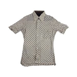 Puede incluir: Una camisa de manga corta con botones y un estampado de lunares negros, marrones y grises sobre un fondo crema. La camisa tiene un cuello marrón oscuro y un bolsillo en el pecho.