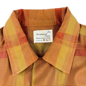 Op de afbeelding: Een bruin en oranje geruit overhemd met een label met de tekst "Permanent Press" en "Never needs ironing", gestyled door "No-Fade".