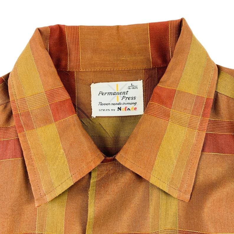 Op de afbeelding: Een bruin en oranje geruit overhemd met een label met de tekst "Permanent Press" en "Never needs ironing", gestyled door "No-Fade".