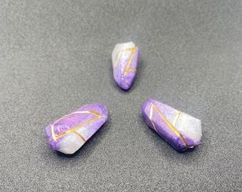 May include: Three purple and white ombre crystal-shaped beads with gold accents. The beads are on a gray surface.