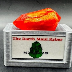 May include: A red and orange Kyber crystal on a silver display stand with the text "The Darth Maul Kyber" and a green Kyber crystal logo.
