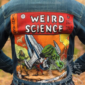 May include: A denim jacket with a comic book style design on the back. The comic book cover features a green dinosaur, a spaceship, and the words "Weird Science" in white letters on a red background.