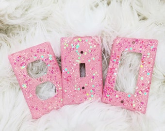 May include: Three pink light switch and outlet covers with glitter. The covers are on a white fluffy surface.