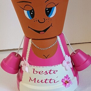 Könnte beinhalten: Ein rosa-weißer Blumentopf mit einem bemalten Gesicht, einer rosa Schleife und den Worten "beste Mutti" auf der Vorderseite. Der Topf hat zwei rosa Glöckchen daran befestigt.