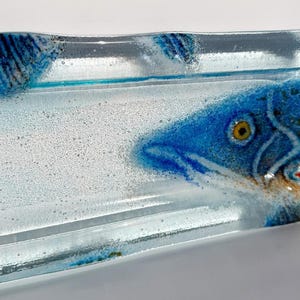 May include: A rectangular glass dish with a fish design. The fish is primarily blue with yellow and red accents. The dish is clear with a textured surface and measures approximately 20 cm long.