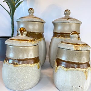 May include: Set of four ceramic canisters with lids. The canisters are brown and white with a speckled glaze. The lids have a brown and white speckled glaze with a brown band around the top.