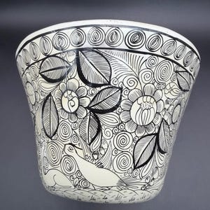 May include: A white ceramic bowl with a black floral and animal design. The bowl features a repeating pattern of flowers, leaves, and swirls, with two stylized seals. The rim has a decorative border.