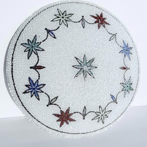 May include: A circular glass lampshade with a textured surface and a floral pattern. The design includes a central star and a border of star-shaped flowers in blue, green, and red, linked by a chain. The edge is outlined with a black line.