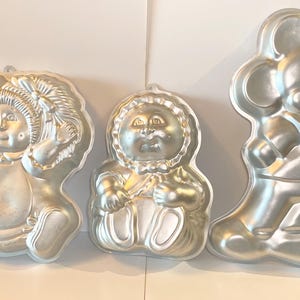 May include: Three silver cake pans, one shaped like a baby doll, one shaped like a girl with pigtails, and one shaped like Mickey Mouse.