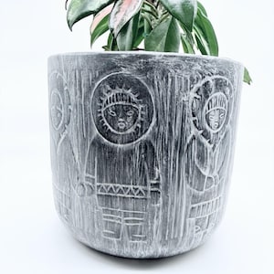 May include: A cylindrical grey planter with a distressed, brushed finish featuring carved relief figures wearing hooded parkas. The planter contains a plant with variegated green and pink leaves extending from the top opening.