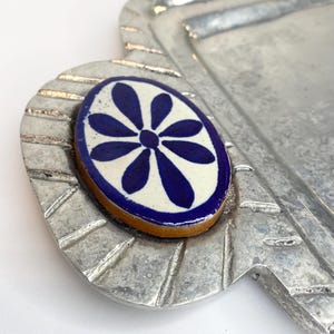 May include: A silver serving tray with a decorative blue and white floral tile on the handle. The tray has a textured surface and a raised edge.