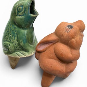 May include: Two ceramic garden stakes featuring animal figures. One is a green glazed frog with a textured back and an open mouth. The other is a matte terracotta-coloured rabbit with painted eyes and ears, both designed with a pointed base for soil.