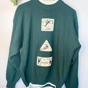 Peut inclure: Un pull en tricot vert foncé avec une bordure côtelée blanche autour du cou et du bas. Le pull présente trois patchs brodés avec des scènes de ski et le texte "St. Moritz", "Rickman 1973" et "Suisse - 1984".