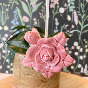 Peut inclure: Ornement en forme de rose en céramique rose, suspendu par un ruban crème. La fleur présente des pétales et des feuilles détaillés. L'ornement est suspendu devant une plante dans un pot en céramique marron, sur fond de papier peint floral.