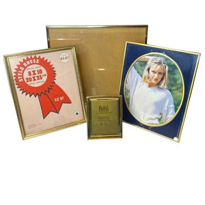 May include: Four gold-coloured picture frames of varying sizes and designs. The largest frame is empty, while the other three frames contain images. One frame has a red and white label with the text "Style House Special Offer 8x10 20x25 cm Offer Speciale Dept 300 $2.87". Another frame has a blue background with a gold oval border and a photo of a woman. The smallest frame has a gold label with the text "Solid Brass Engraved Solid Brass Caption Holder 4" x 6" Made in Italy".