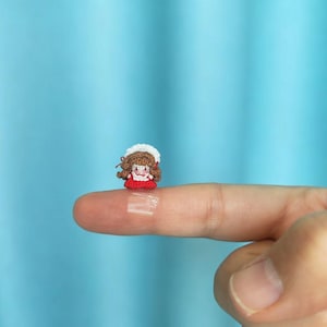 以下が含まれることがあります： 茶色の糸の髪、白いボンネット、赤いドレスを特徴とするミニチュアのかぎ針編みの人形。この小さな手作りのキャラクターは細い糸で構成されており、顔と衣服に複雑なステッチのディテールが施されています。