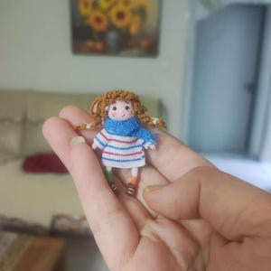 以下が含まれることがあります： 茶色の髪の毛をした小さな編みぐるみ人形。青いマフラーと白と青のストライプのシャツを着ています。人形は小さく、青と白のストライプのバッグを持っています。