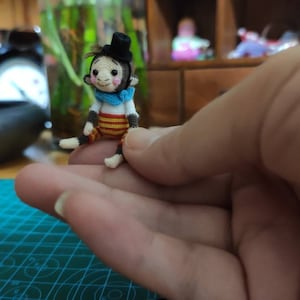 May include: A small crocheted monkey wearing a black top hat and a blue and white striped shirt. The monkey is sitting on a person's hand.