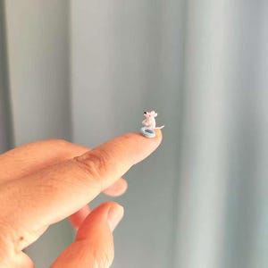 May include: A miniature crocheted white mouse figurine sitting on a small, light blue crocheted ring. The tiny mouse has a pointed snout, small ears, and a thin tail, resting on the circular base, showcasing intricate needlework and fine detail.