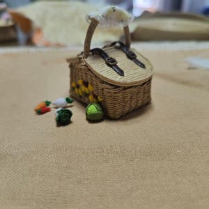 以下が含まれることがあります： ハンドルと2本の革ストラップで固定された蓋が付いた、ミニチュアの織りピクニックバスケット。バスケットは黄色のヒマワリのアクセントで装飾されています。ニンジン、キャベツ、緑色のボールなど、小さなかぎ針編みの野菜が近くに散らばっています。