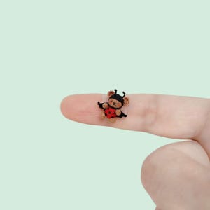 May include: A miniature crocheted teddy bear figurine wearing a black and red ladybug costume. The tiny bear has light brown yarn fur, black antennae, and a red body with black spots. It is crafted with fine thread and intricate stitching details.