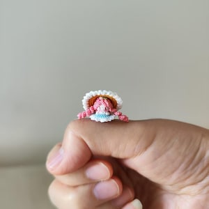 May include: A miniature crochet doll with a pink head, arms, and legs, wearing a white hat with a yellow brim and a blue and white dress. The doll is resting on a person's finger. The doll is handmade.