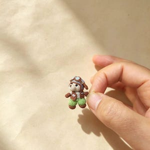May include: A miniature crocheted bear figurine wearing a brown and light blue aviator hat with goggles, a brown vest, and green trousers. The small, detailed stuffed animal is crafted with fine yarn and features a light tan face and ears.