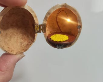 May include: A miniature, hinged walnut shell opens to reveal a small, illuminated interior. Inside, a small, round, brown platform holds a yellow, textured object. The shell is held open by a small brass hinge.