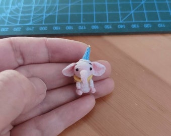 May include: A small pink crocheted elephant wearing a blue party hat. The elephant has yellow yarn for its eyes and mouth.